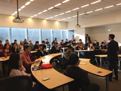 Students and faculty seated in a tiered classroom at the kickoff meeting, with a speaker standing at the right.