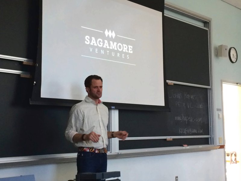 The speaker standing at the front of the room while students and guests sit at tables.